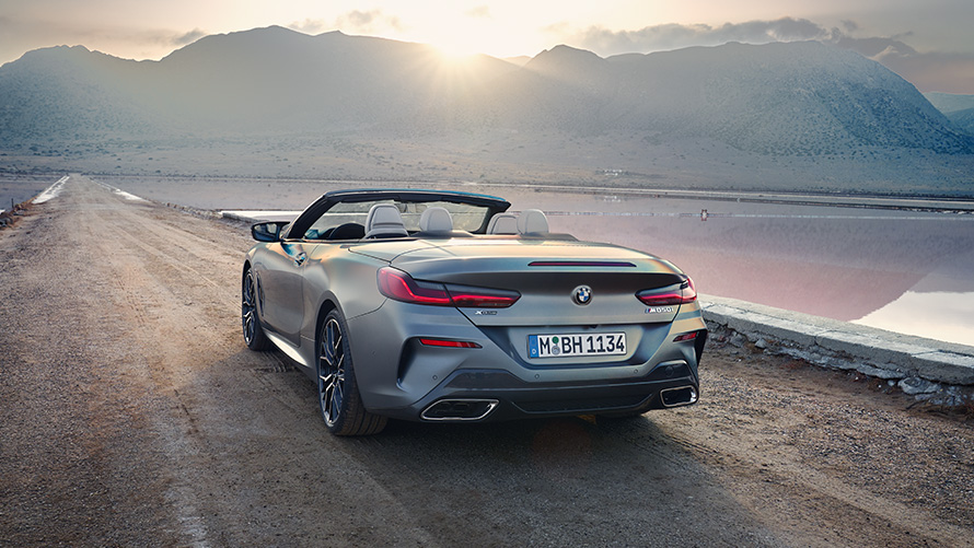 The new BMW 8 Series Convertible G14 LCI Facelift 2022 BMW Individual Frozen Pure Grey metallic rear view with open softtop standing on sandy road with hills in the background