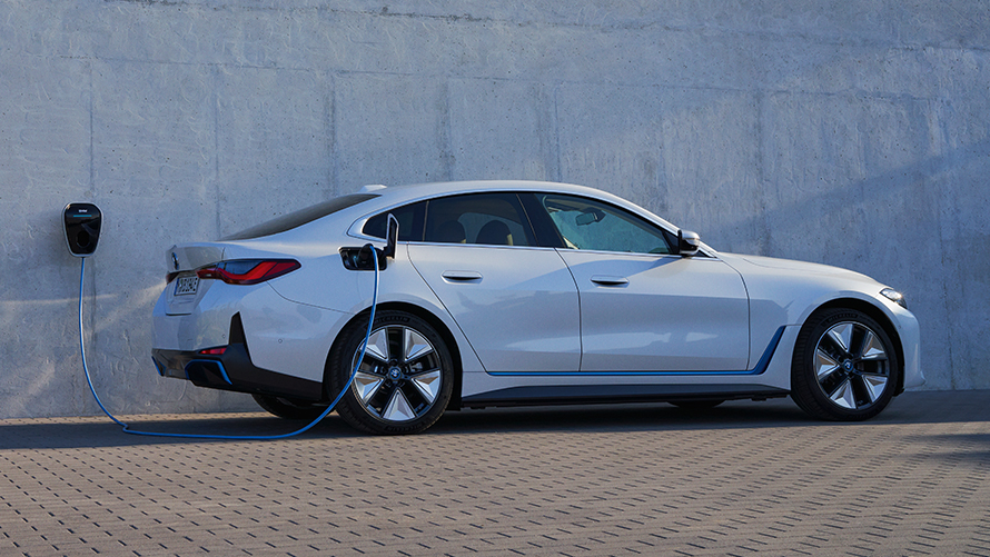 Vue de profil avec la BMW Wallbox BMW i4 eDrive40 G26 2021 BMW i4 eDrive40 G26 2021 Mineralweiß Vue de profil devant un mur avec une borne de recharge BMW Wallbox