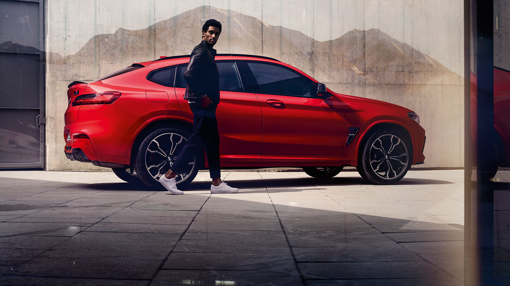 BMW X4 M Competition, side view with young man. BMW X4 M Competition in Toronto Red metallic, exterior, side view with young man.