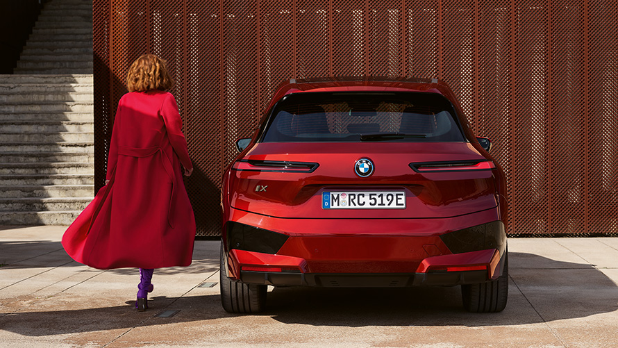Woman in red coat next to BMW iX (i20) closing the tailgate Closing the tailgate Woman in red coat next to BMW iX (i20) in front of building