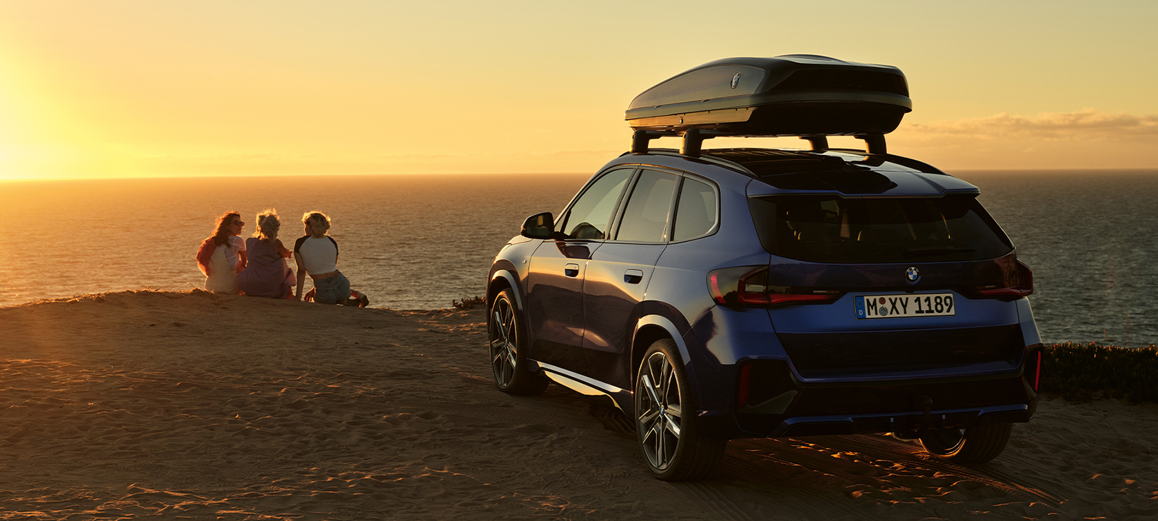 Original BMW Accessories for Spring and Summer BMW X1 U11 Original BMW Accessories for Spring and Summer BMW X1 U11 standing on sand in front of a coast with 2 people